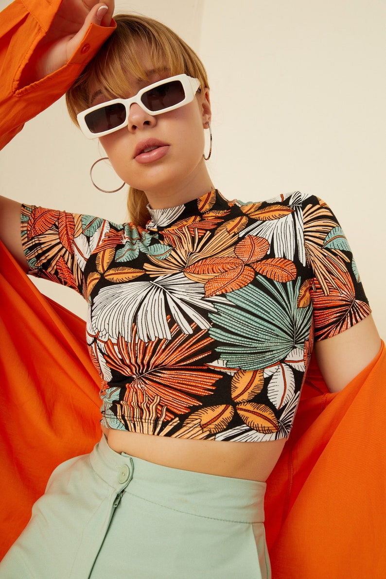May include: A black crop top with a tropical leaf print. The top has a mock neck and short sleeves. The model is wearing the top with a pair of light green pants and a bright orange shirt.