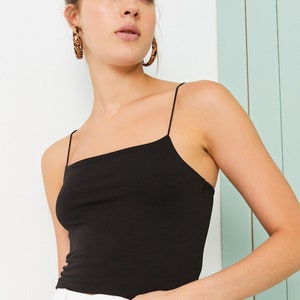 May include: A black spaghetti strap tank top with a square neckline. The model is wearing white jeans and has on large hoop earrings.