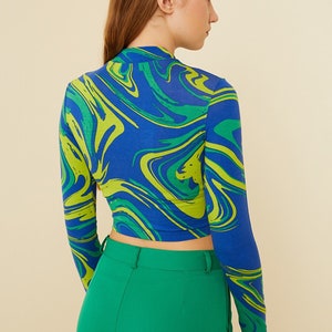 Pu&ograve; includere: Un top corto a maniche lunghe blu e verde con una stampa astratta a spirale. Il top &egrave; indossato con pantaloni verdi a vita alta.