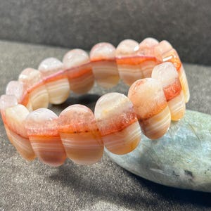 Puede incluir: Un brazalete hecho de piedras de ágata bandeada. Las piedras son una mezcla de colores blanco, rosa y naranja. El brazalete está hecho de piedras lisas y redondeadas.