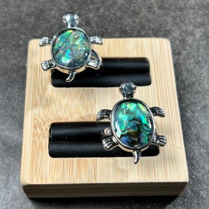 May include: Two silver-toned rings with turtle designs. Each ring features an oval-shaped abalone shell center with iridescent green and blue hues. The rings are displayed on a wooden jewelry holder with black ring supports.