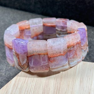 Puede incluir: Un brazalete hecho de cuentas de ágata en capas en tonos de rosa, morado y naranja. Las cuentas son rectangulares y tienen un acabado pulido.