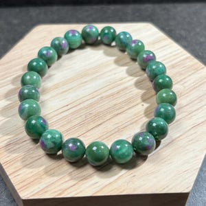 May include: A green beaded bracelet with red and purple inclusions. The round beads are strung together, creating a circular shape. The bracelet is displayed on a wooden surface.