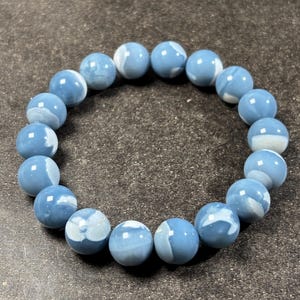 Könnte beinhalten: Ein Armband mit runden, hellblauen Perlen mit weißen Marmorierungen. Die Perlen sind aneinandergereiht und bilden eine kreisförmige Form. Das Armband besteht aus polierten Steinen und hat eine weiche, pastellblaue Farbe.