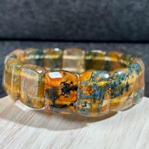 May include: A gemstone bracelet featuring rectangular beads with a mix of orange, yellow, and blue-green patterns. The bracelet is displayed on a wooden surface, showcasing the natural variations in the stone.