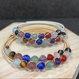 May include: Two bracelets with colorful gemstone beads. The bracelets have gold and silver-colored metal bands. The beads are various colors, including blue, green, red, and purple. The bracelets are displayed on a wooden surface.