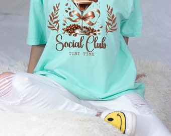 May include: A turquoise blue t-shirt with a graphic of a martini glass with coffee beans and a brown ribbon. The text "Espresso Martini Social Club Tini Time" is printed on the shirt.