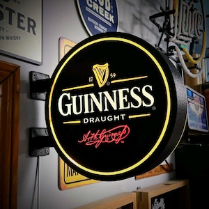May include: A circular, wall-mounted, illuminated Guinness Draught sign with a black background, a gold harp logo, white lettering, and a red Arthur Guinness signature. The sign features a glowing yellow border and a black metal mounting bracket.