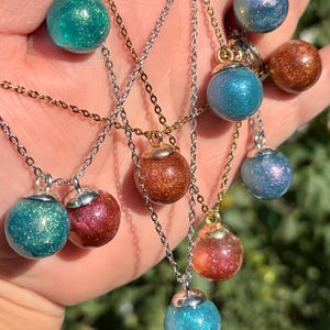 May include: A collection of glitter globe necklaces in various colors, including teal, red, blue, and copper. Each necklace features a small, round globe pendant suspended from a delicate chain. The pendants have a metallic cap.