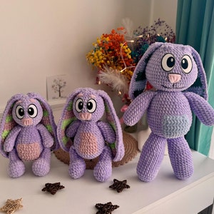 May include: Three crocheted purple bunny plushies with pink noses and green accents. The largest bunny is standing, while the two smaller bunnies are sitting.