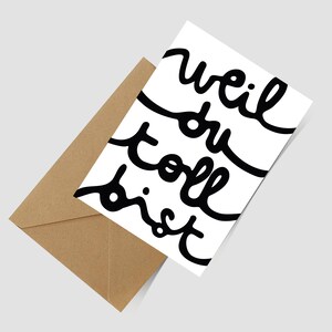 May include: A white greeting card with black handwritten cursive text that reads "weil du toll bist" positioned diagonally next to a brown kraft paper envelope. The card features a clean design with bold, flowing lettering across the front surface.