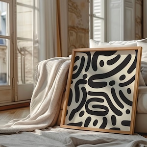 Puede incluir: Una impresión abstracta enmarcada con un patrón blanco y negro de líneas curvas y puntos. La impresión se apoya en un sofá blanco con una manta blanca.