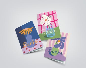 May include: Three folded greeting cards featuring floral illustrations and the word Danke in cursive script. One card shows an orange flower in a blue vase on a polka dot surface, another a white flower in a blue vase on a grid, and one a purple flower.