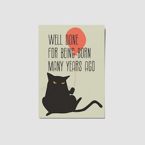 May include: A rectangular greeting card with a light beige background featuring a black cat with yellow eyes holding a red balloon. The text reads: WELL DONE FOR BEING BORN MANY YEARS AGO. The cat is depicted in a seated position with its tail extended.