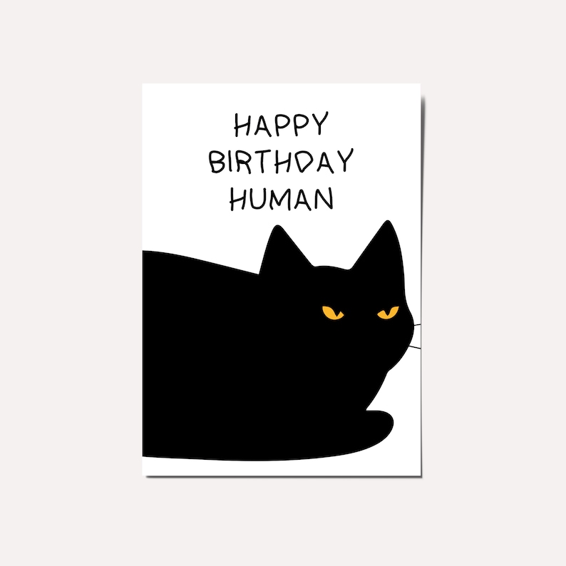 May include: A black cat with yellow eyes peeking over a white card with the text "Happy Birthday Human" written in black.