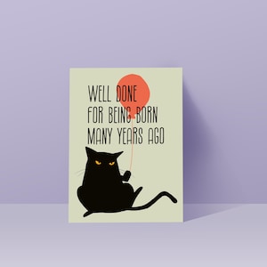 May include: A rectangular greeting card with a light green background featuring a black cat with yellow eyes holding a red balloon. The text printed in black reads, Well done for being born many years ago. The card has a clean, graphic design.