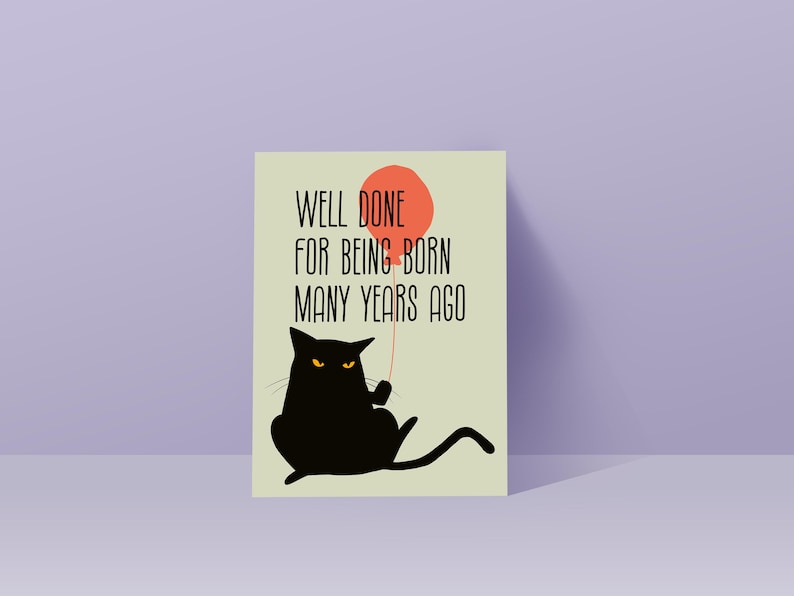 May include: A rectangular greeting card with a light green background featuring a black cat with yellow eyes holding a red balloon. The text printed in black reads, Well done for being born many years ago. The card has a clean, graphic design.