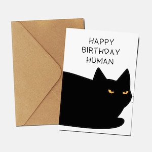 May include: A white card with black text that says "Happy Birthday Human" and a black cat with yellow eyes peeking out from the bottom of the card.