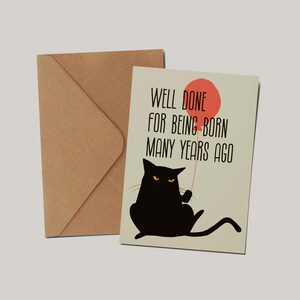 May include: A greeting card with a light beige background featuring a black cat holding a red balloon and the text WELL DONE FOR BEING BORN MANY YEARS AGO in black font, accompanied by a plain brown kraft paper envelope positioned behind it.