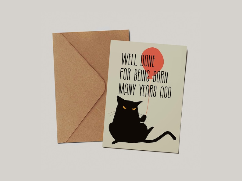May include: A greeting card with a light beige background featuring a black cat holding a red balloon and the text WELL DONE FOR BEING BORN MANY YEARS AGO in black font, accompanied by a plain brown kraft paper envelope positioned behind it.
