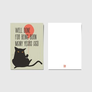 May include: A rectangular greeting card featuring a black cat with orange eyes holding a red balloon. The text reads, "WELL DONE FOR BEING BORN MANY YEARS AGO." The card is shown from the front and the back, with a small red logo on the back side.