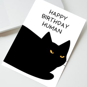 May include: A black cat with yellow eyes peeks over a white card with the text "Happy Birthday Human" written in black.