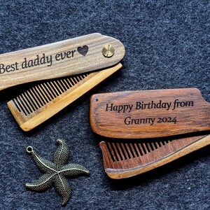 K&ouml;nnte beinhalten: Zwei Holz-Klapptaschenkamm mit gravierten Botschaften. Ein Kamm sagt "Best daddy ever" mit einem Herzsymbol. Der andere Kamm sagt "Happy Birthday from Granny 2024".