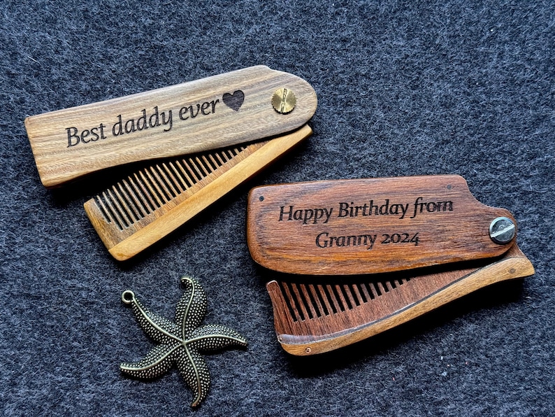 K&ouml;nnte beinhalten: Zwei Holz-Klapptaschenkamm mit gravierten Botschaften. Ein Kamm sagt "Best daddy ever" mit einem Herzsymbol. Der andere Kamm sagt "Happy Birthday from Granny 2024".