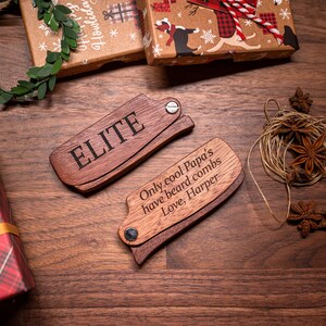 K&ouml;nnte beinhalten: Zwei Holz-Bartscheider mit einer personalisierten Nachricht. Der obere Kamm sagt "ELITE" und der untere Kamm sagt "Nur coole Papas haben Bartscheider. Liebe, Harper".