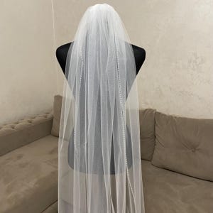 May include: A white bridal veil with a beaded trim. The veil is long and flowing, and it is displayed on a mannequin.