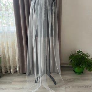 Puede incluir: Un velo de novia blanco de una sola capa de tul con un dise&ntilde;o de cuentas. El velo se muestra en un maniqu&iacute; y cuelga hasta el suelo.