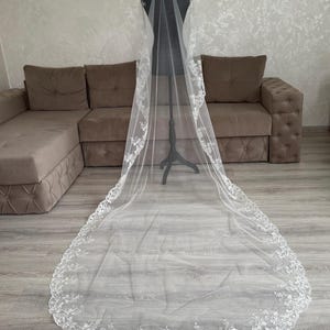 K&ouml;nnte beinhalten: Ein langer, wei&szlig;er Brautschleier mit aufwendigen floralen Spitzenverzierungen an den R&auml;ndern. Der Schleier wird auf einer Schaufensterpuppe pr&auml;sentiert, die seine L&auml;nge und sein zartes Design zeigt. Aus leichtem, transparentem Stoff.