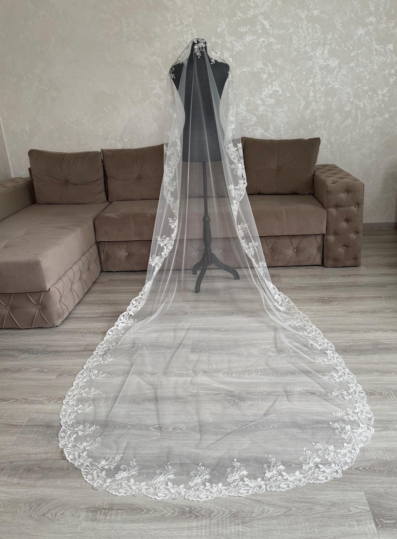 K&ouml;nnte beinhalten: Ein langer, wei&szlig;er Brautschleier mit aufwendigen floralen Spitzenverzierungen an den R&auml;ndern. Der Schleier wird auf einer Schaufensterpuppe pr&auml;sentiert, die seine L&auml;nge und sein zartes Design zeigt. Aus leichtem, transparentem Stoff.