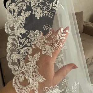 K&ouml;nnte beinhalten: Ein wei&szlig;er Brautschleier mit aufwendigen floralen Spitzendetails. Die Spitze weist ein sich wiederholendes Muster aus Blumen und Bl&auml;ttern auf, das einen zarten und eleganten Rand bildet. Der Schleier ist durchscheinend, l&auml;sst Licht durch und besteht wahrscheinlich aus T&uuml;ll oder einem &auml;hnlichen Stoff.