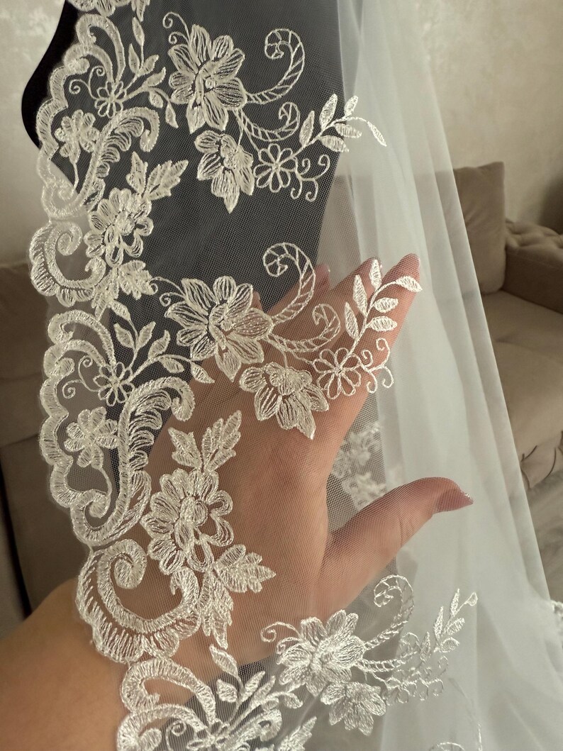 K&ouml;nnte beinhalten: Ein wei&szlig;er Brautschleier mit aufwendigen floralen Spitzendetails. Die Spitze weist ein sich wiederholendes Muster aus Blumen und Bl&auml;ttern auf, das einen zarten und eleganten Rand bildet. Der Schleier ist durchscheinend, l&auml;sst Licht durch und besteht wahrscheinlich aus T&uuml;ll oder einem &auml;hnlichen Stoff.