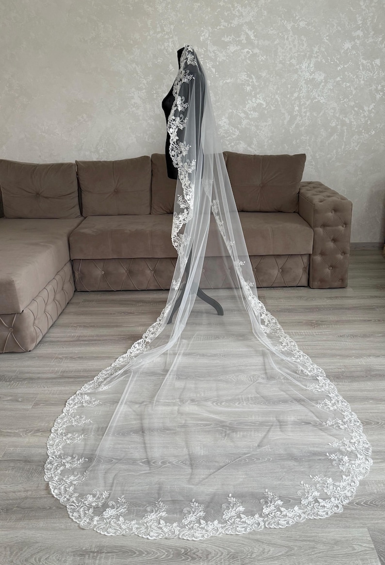 K&ouml;nnte beinhalten: Ein langer, wei&szlig;er Brautschleier mit aufwendigen floralen Spitzenverzierungen an den R&auml;ndern. Der Schleier wird auf einer schwarzen Schaufensterpuppe pr&auml;sentiert, die seine L&auml;nge und sein zartes Design zeigt. Der Schleier besteht aus einem transparenten, leichten Stoff.