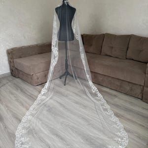 K&ouml;nnte beinhalten: Ein langer, wei&szlig;er Brautschleier mit aufwendigen floralen Spitzenverzierungen an den R&auml;ndern. Der Schleier wird auf einer schwarzen Schaufensterpuppe pr&auml;sentiert, die seine L&auml;nge und sein zartes Design zeigt. Der transparente Stoff l&auml;sst Licht durchscheinen und hebt die Muster der Spitze hervor.