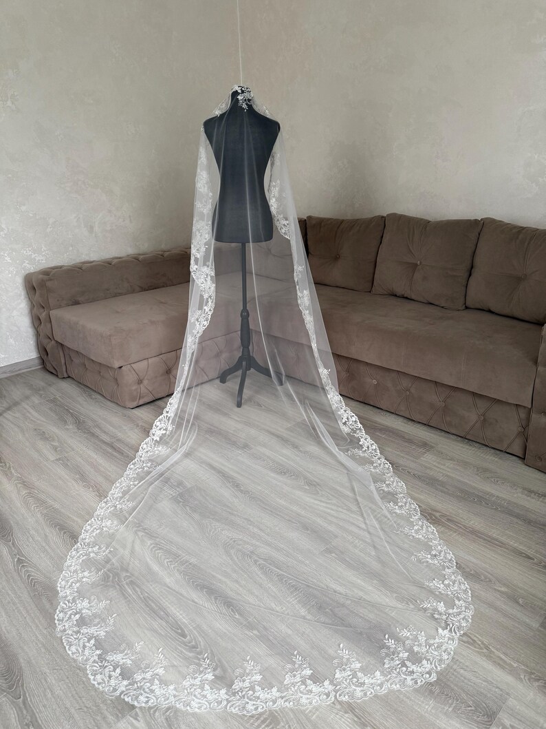 K&ouml;nnte beinhalten: Ein langer, wei&szlig;er Brautschleier mit aufwendigen floralen Spitzenverzierungen an den R&auml;ndern. Der Schleier wird auf einer schwarzen Schaufensterpuppe pr&auml;sentiert, die seine L&auml;nge und sein zartes Design zeigt. Der transparente Stoff l&auml;sst Licht durchscheinen und hebt die Muster der Spitze hervor.