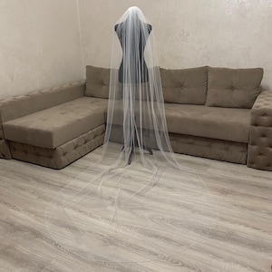 May include: A long, white tulle wedding veil draped over a dress form. The veil is very sheer and has a delicate, flowing design.