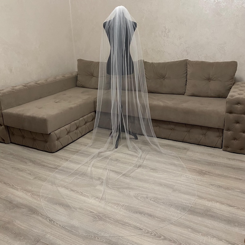 May include: A long, white tulle wedding veil draped over a dress form. The veil is very sheer and has a delicate, flowing design.