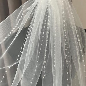 Puede incluir: Un velo de novia blanco de una sola capa de tul con un borde de cuentas. El velo est&aacute; decorado con peque&ntilde;as cuentas espaciadas uniformemente.