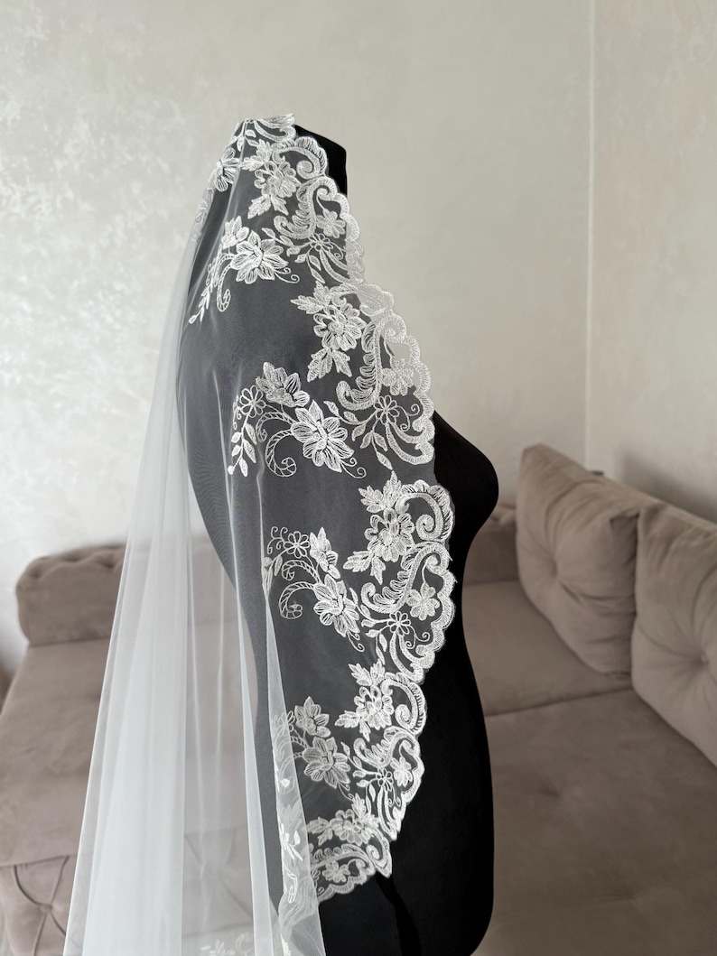 K&ouml;nnte beinhalten: Ein wei&szlig;er Brautschleier mit zarter floraler Spitzenborte. Die Spitze weist aufwendige Designs von Blumen und Bl&auml;ttern auf, die einen Hauch von Eleganz verleihen. Der Schleier ist durchsichtig und f&auml;llt anmutig.