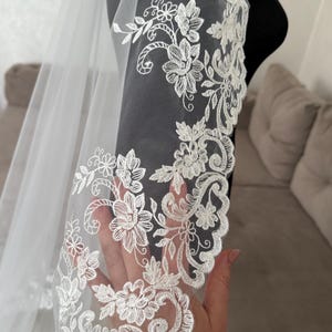 K&ouml;nnte beinhalten: Ein wei&szlig;er Brautschleier mit aufwendigen floralen Spitzenverzierungen am Rand. Die zarte Spitze weist gestickte Blumen und wirbelnde Muster auf, die einen eleganten und romantischen Look erzeugen. Der Schleier ist durchsichtig und leicht.