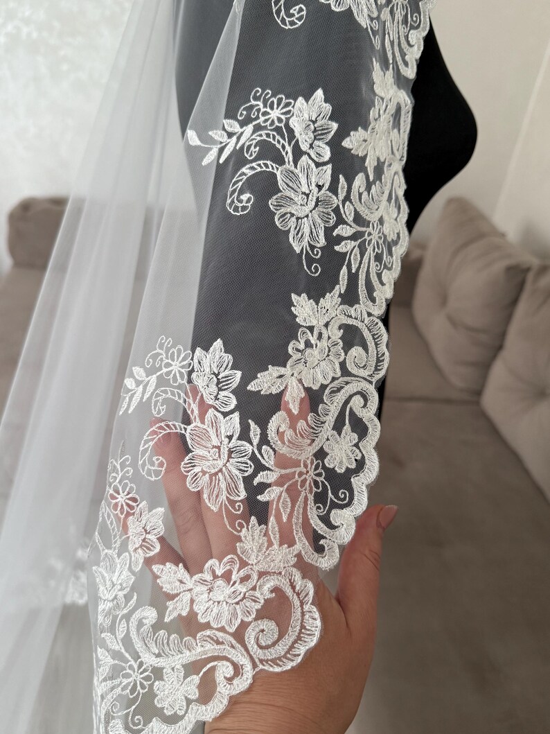 K&ouml;nnte beinhalten: Ein wei&szlig;er Brautschleier mit aufwendigen floralen Spitzenverzierungen am Rand. Die zarte Spitze weist gestickte Blumen und wirbelnde Muster auf, die einen eleganten und romantischen Look erzeugen. Der Schleier ist durchsichtig und leicht.
