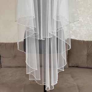 May include: A white, layered bridal veil with a beaded edge. The veil is displayed on a mannequin, showcasing its delicate, flowing design. The beads add a subtle sparkle, enhancing the veil's elegance. The background features a neutral-toned sofa.