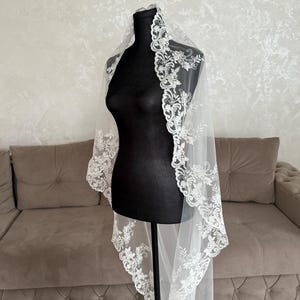 K&ouml;nnte beinhalten: Ein wei&szlig;er Brautschleier mit aufwendigen floralen Spitzendetails an den R&auml;ndern. Der Schleier ist &uuml;ber einer schwarzen Schaufensterpuppe drapiert, was sein zartes Design und seinen transparenten Stoff hervorhebt. Im Hintergrund ein Sofa und eine Wand in neutralen T&ouml;nen.