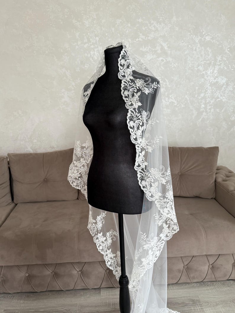 K&ouml;nnte beinhalten: Ein wei&szlig;er Brautschleier mit aufwendigen floralen Spitzendetails an den R&auml;ndern. Der Schleier ist &uuml;ber einer schwarzen Schaufensterpuppe drapiert, was sein zartes Design und seinen transparenten Stoff hervorhebt. Im Hintergrund ein Sofa und eine Wand in neutralen T&ouml;nen.
