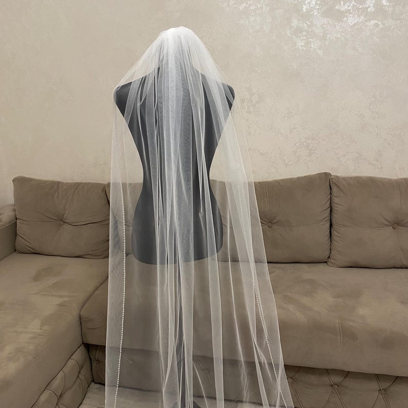 May include: A white bridal veil with a beaded edge, displayed on a mannequin. The veil is long and flowing, with a delicate, sheer fabric.