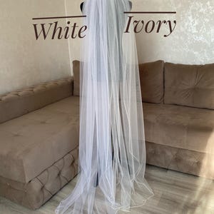 May include: A white bridal veil with a beaded edge. The veil is draped over a brown couch with the words "White" and "Ivory" written on the wall behind it.