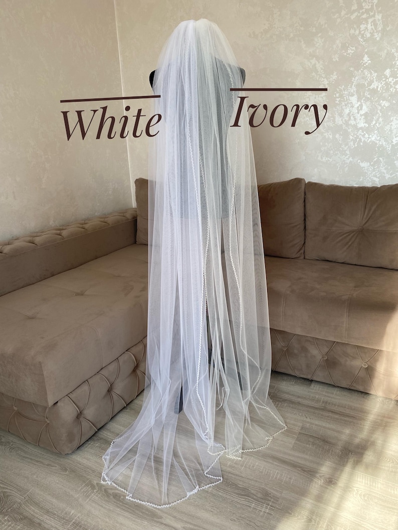 May include: A white bridal veil with a beaded edge. The veil is draped over a brown couch with the words "White" and "Ivory" written on the wall behind it.